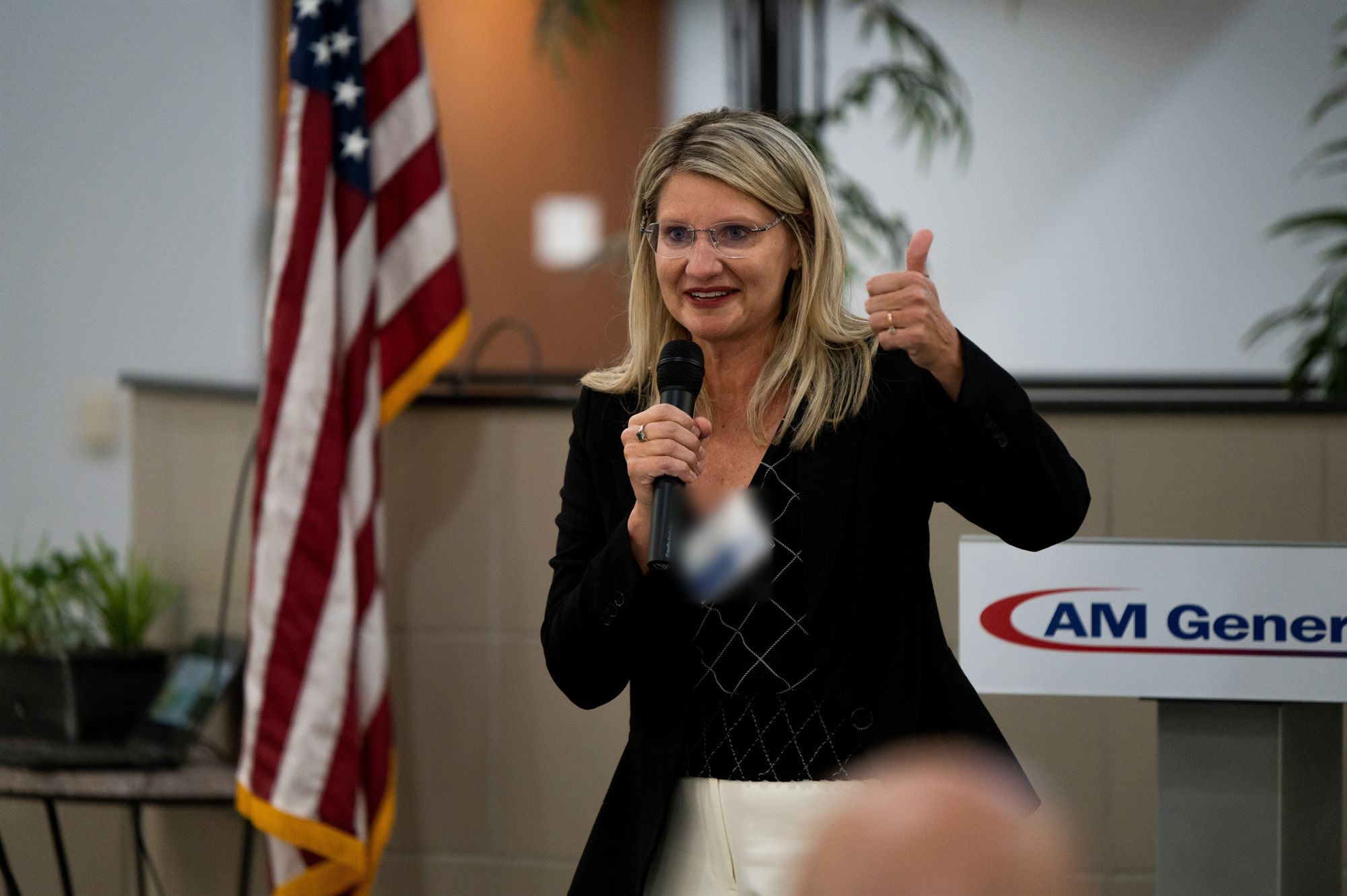 Cheryl Thompson speaking into a microphone at a manufacturing industry event, standing beside a United States flag.