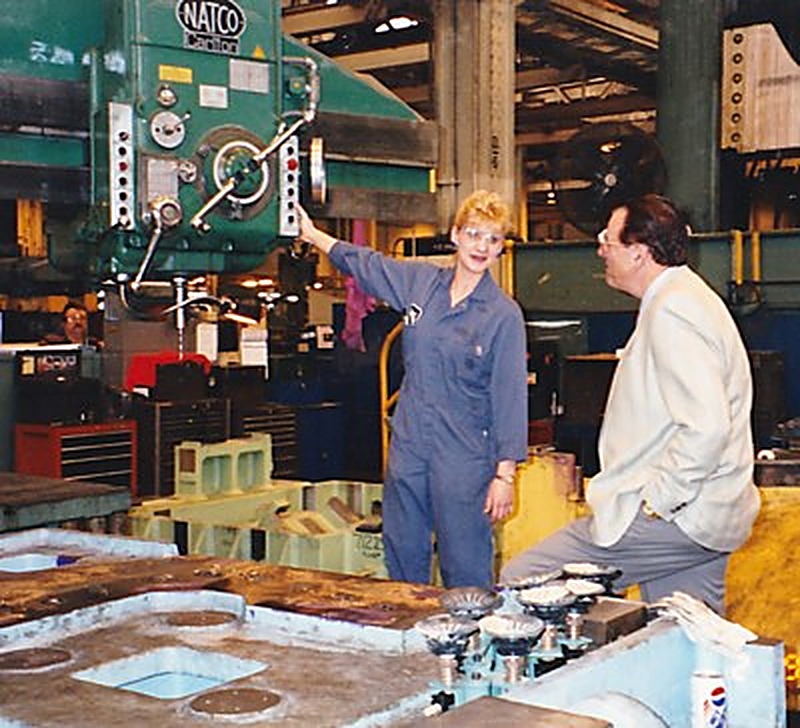 Cheryl Thompson in coveralls and safety glasses at a large press on the tool and die floor, talking with a manager.
